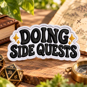 May include: A white embroidered patch with a scalloped edge featuring the black text DOING SIDE QUESTS in a bold, rounded font. The design includes four small, four-pointed gold stars placed around the lettering on the white fabric.