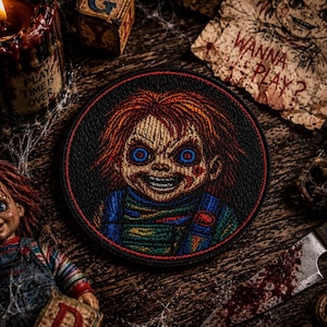 May include: A circular embroidered patch featuring a detailed portrait of a doll with messy orange hair, bright blue eyes, and a wide, toothy grin. The doll wears a blue overall-style garment with colorful striped sleeves and a red circular accent.
