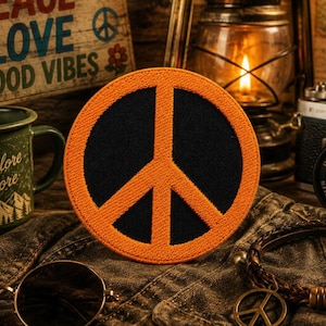 May include: A round, orange and black peace sign patch. The patch is on a denim jacket, with a vintage aesthetic. Other items in the image include a green mug, sunglasses, and a leather bracelet with a peace sign charm.