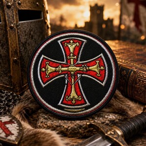 May include: Round black patch with a red cross design, outlined in white and gold. The patch is embroidered with a detailed medieval cross. The background includes a medieval helmet, a sword, and a shield.
