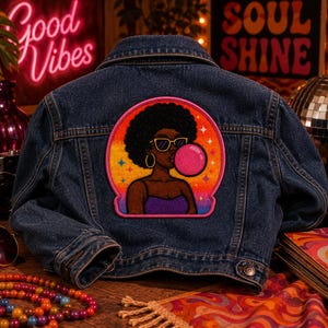 Pode incluir: Um casaco de ganga azul escuro com um grande emblema bordado nas costas. O emblema retrata uma pessoa com penteado afro, &oacute;culos de sol amarelos e brincos de argola, a soprar uma pastilha el&aacute;stica cor-de-rosa contra um p&ocirc;r do sol.