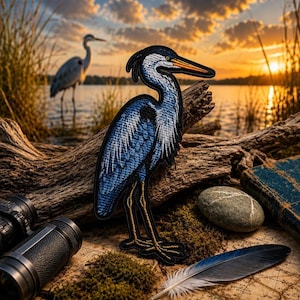 May include: Embroidered patch of a blue heron with a black outline, perched on a log. The patch features detailed stitching and a vibrant color palette. The background includes a scenic view of a lake at sunset, binoculars, a book, and a feather.