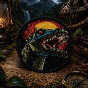 May include: Circular embroidered patch depicting a dinosaur head with an open mouth, set against a yellow sun and red sky. The dinosaur is blue and green with red eyes. The patch has a black border, suitable for clothing or accessories.