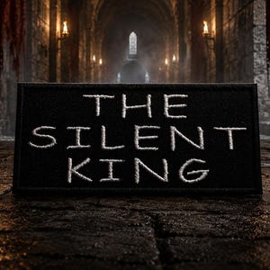 Pode incluir: Um patch retangular preto com as palavras "THE SILENT KING" bordadas em branco. O patch está situado contra um fundo escuro e atmosférico de um corredor de pedra com tochas e uma luz distante.