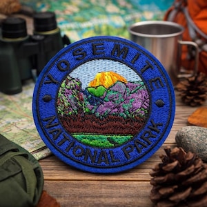 以下が含まれることがあります： 鮮やかな山岳風景を囲むように「YOSEMITE NATIONAL PARK」の文字が刺繍された、丸い青色のパッチ。黄色とオレンジ色の山、紫と緑の木々、緑色の湖が描かれています。パッチは木製の表面に置かれています。