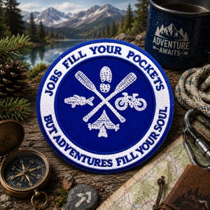 Peut inclure: Un écusson brodé bleu circulaire avec une bordure blanche portant le texte JOBS FILL YOUR POCKETS BUT ADVENTURES FILL YOUR SOUL. Le centre présente des icônes blanches brodées de rames croisées, une empreinte, un poisson, un vélo et une tente.