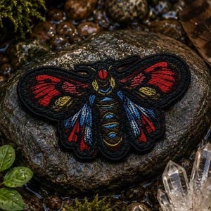 Op de afbeelding: Een geborduurde patch in de vorm van een nachtvlinder met een zwarte rand. De vlinder heeft een rode kop, gele en blauwe accenten op het borststuk en vleugels met patronen van felrood, blauw en geel garen op een zwarte achtergrond.