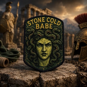 May include: A rectangular embroidered patch with a black border featuring a green-toned illustration of a face with snakes for hair. The text "STONE COLD BABE" is stitched in yellow thread at the top of the patch against a dark green background.