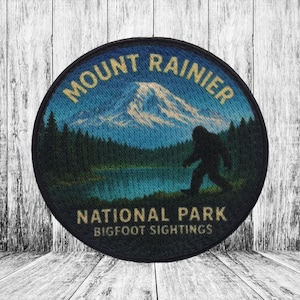 Puede incluir: Parche redondo con el texto "Mount Rainier National Park Bigfoot Sightings". El diseño incluye una montaña, un lago, árboles y una silueta de Bigfoot. El parche tiene un borde negro. Los colores son azul, verde y beige.