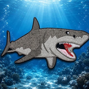Puede incluir: Parche bordado con un tiburón gris y blanco detallado con la boca abierta que revela dientes rojos y blancos. El parche tiene una apariencia texturizada y está colocado sobre un fondo submarino azul con burbujas y coral.