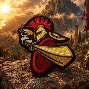 Könnte beinhalten: Ein gestickter Aufnäher mit einem goldenen spartanischen Helm mit rotem Kamm, platziert über einem goldenen Schwert und einem runden roten Schild. Das Design ist mit schwarzem Faden umrandet und zeigt detaillierte Stickereien der Rüstung.