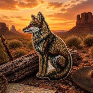 Op de afbeelding: Een geborduurde patch met een zittende coyote, uitgevoerd in bruine, beige en witte draden. De coyote is in profiel naar links afgebeeld, met gedetailleerd stikwerk dat de textuur van de vacht en de puntige oren van het dier benadrukt.