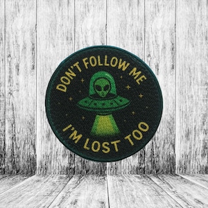 May include: Round black patch with the text "DON'T FOLLOW ME I'M LOST TOO" in gold lettering. The patch features a green alien in a UFO with a green beam of light. The patch is on a white and gray wooden background.