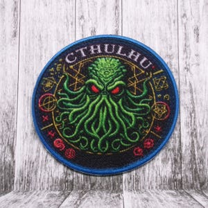 May include: A round, blue-edged patch featuring a green, tentacled creature with red eyes at its centre. The creature is encircled by a gold and black circular design with the word "CTHULHU" in a gold font.