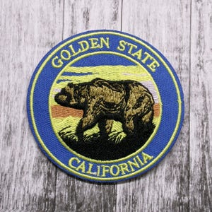 Puede incluir: Un parche bordado azul y amarillo con un oso marrón caminando sobre un fondo de puesta de sol. El texto "Golden State California" está bordado alrededor del oso.