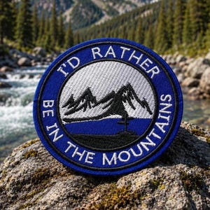 Peut inclure: Écusson brodé rond, bleu et blanc, avec le texte "I'D RATHER BE IN THE MOUNTAINS". Le motif représente une chaîne de montagnes en noir et blanc. L'écusson est placé sur un fond extérieur naturel.