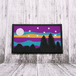 May include: An embroidered patch with a purple, blue, and yellow sunset scene. The patch features a white moon, stars, and a silhouette of trees and mountains.