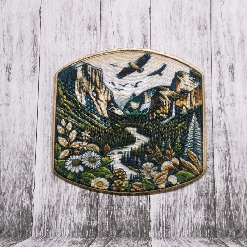 May include: Embroidered patch featuring a scenic mountain landscape with a river, trees, and flying birds. The patch has a gold border and includes detailed depictions of flowers and foliage in shades of green, brown, and white.