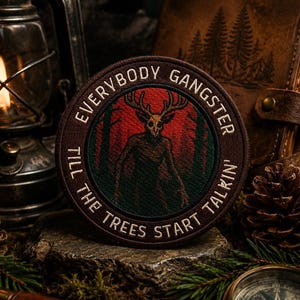 以下が含まれることがあります： 「EVERYBODY GANGSTER TILL THE TREES START TALKIN'」の文字が入った丸い茶色のパッチ。鹿の頭蓋骨を持つ生き物が、赤い背景の森の中に描かれています。