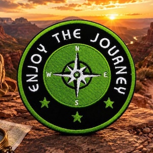 May include: A circular embroidered patch featuring a black border with the white text "ENJOY THE JOURNEY" and three green stars. The center contains a green circle with a white compass rose design marked with N, S, E, and W directional letters.