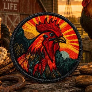 May include: A circular embroidered patch featuring a detailed rooster head in vibrant red, orange, and yellow tones. The design includes a sunburst background with rays extending outward, set against dark green mountains and a light blue circular moon.