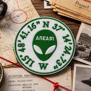 May include: A circular embroidered patch featuring a green alien head silhouette in the center with the text AREA 51 above it. The white border contains the embroidered coordinates 37 14 24.73 N and 115 48 41.16 W in green thread.