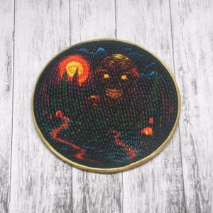Puede incluir: Un parche redondo bordado con una figura oscura y sombría con ojos rojos brillantes y una sonrisa amenazante. La figura se coloca sobre un fondo oscuro con una luna naranja brillante y un bosque en la distancia.