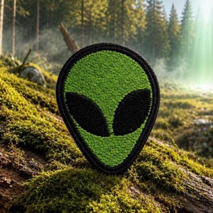 May include: An embroidered patch in the shape of an alien head, featuring a vibrant lime green face with two large, solid black oval eyes, all outlined by a thick, dark black stitched border that defines the distinct teardrop-like silhouette.