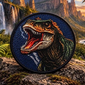 May include: A circular embroidered patch featuring a detailed depiction of a Tyrannosaurus Rex with an open mouth, sharp teeth, and a textured hide in shades of orange, green, and brown against a dark blue background with a black border.