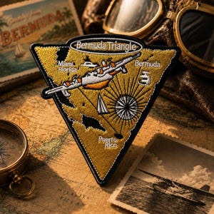 May include: Embroidered patch in a triangular shape with a gold background and black border. The patch features the text "Bermuda Triangle" and illustrations of an airplane, a ship, and a compass. The patch also includes the text "Miami Florida", "Bermuda", and "Puerto Rico".