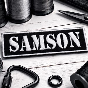 May include: A rectangular black embroidered patch with the white text SAMSON in a bold serif font. The patch is surrounded by black and grey sewing thread spools, black metal scissors, a black carabiner, a black ring, and a measuring tape.