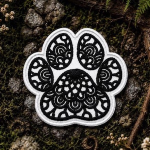 May include: A white embroidered patch in the shape of a paw print featuring intricate black thread detailing. The design consists of a large central pad and four smaller toe pads, all filled with detailed, swirling, and scalloped black embroidery.