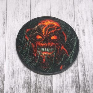 Puede incluir: Un parche redondo con borde negro que presenta un diseño de calavera naranja fuego. La calavera tiene ojos rojos brillantes y una sonrisa amenazante. El fondo es verde oscuro con patrones en espiral.