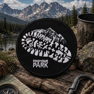 May include: A circular black embroidered patch featuring a white stitched design of a boot print. Inside the boot print are silhouettes of mountains, trees, and a bison. The text "EXPLORE" is stitched above and "FIND YOUR PARK" below.