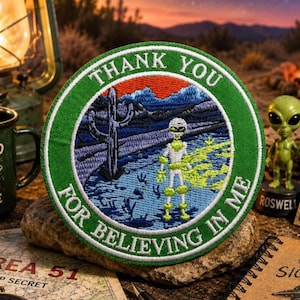 May include: A circular embroidered patch with a green border featuring the text THANK YOU FOR BELIEVING IN ME. The center depicts a white alien figure standing in a desert landscape with a tall cactus, blue mountains, and a bright orange sky.
