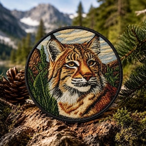 May include: Round embroidered patch featuring a detailed portrait of a bobcat. The patch showcases the animal's orange and brown fur, green foliage, and a mountain backdrop. The patch has a black border.