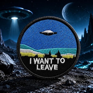 Op de afbeelding: Ronde geborduurde patch met een zwarte rand met een landschap met een UFO en de tekst "I WANT TO LEAVE". De patch heeft een blauwe lucht, groene en gele heuvels en zwarte bomen.