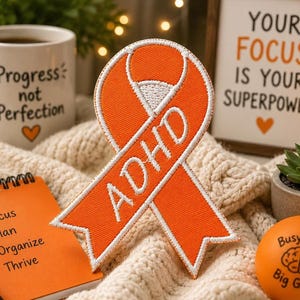 May include: An orange ADHD awareness ribbon patch with white embroidered text and border. A white mug with "Progress not Perfection" and a small orange heart is visible. A small notepad with a checklist and the words "Focus, Plan, Organize, Thrive" is also present.
