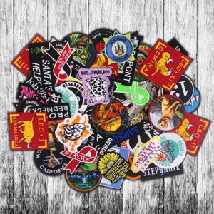 May include: A collection of assorted embroidered patches with various designs and text. The patches feature images of a zombie, a dragon, a sunflower, and text such as "Santa's Help Job 38:4" and "Black Lives Matter". The patches are arranged on a white wooden surface.