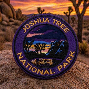 May include: A circular embroidered patch featuring the text "JOSHUA TREE NATIONAL PARK" in gold lettering on a purple background. The patch depicts a desert landscape with Joshua trees and a sunset. The patch is likely a souvenir or collectible item.