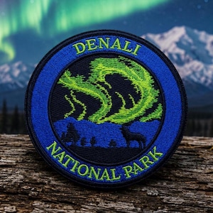 May include: A circular embroidered patch featuring a blue border with the text DENALI NATIONAL PARK in yellow. The center depicts a black silhouette of a wolf standing before blue mountains under a vibrant green aurora borealis night sky.