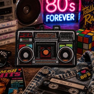 May include: Embroidered patch featuring a retro boombox design with colorful accents. The patch is black with red, orange, yellow, green, and blue details. The background includes a neon sign that says "80's FOREVER", a Rubik's Cube, and cassette tapes.