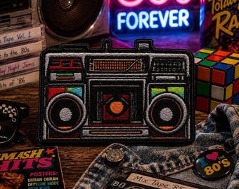 May include: Embroidered patch featuring a retro boombox design with colorful accents. The patch is black with red, orange, yellow, green, and blue details. The background includes a neon sign that says "80's FOREVER", a Rubik's Cube, and cassette tapes.