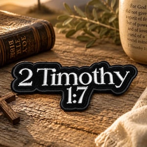 May include: A black and white embroidered patch with the text "2 Timothy 1:7" in a stylized font. The patch is on a wooden surface, with a Bible and a small wooden cross in the background.