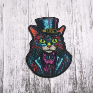 May include: A colorful embroidered patch of a cat wearing a top hat and bow tie. The cat has green eyes and a black nose. The patch is on a white wooden background.