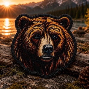 Può includere: Una toppa ricamata circolare che presenta il ritratto dettagliato di un orso bruno con pelliccia marrone dorato, occhi scuri e naso nero. La toppa ha un bordo spesso cucito in nero e mostra un intricato lavoro di filo per i tratti del viso.