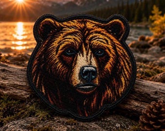 May include: A circular embroidered patch featuring a detailed portrait of a brown bear with golden-brown fur, dark eyes, and a black nose. The patch has a thick black stitched border and showcases intricate thread work to define the bear's facial features.