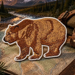 Puede incluir: Un parche bordado con la forma de un oso pardo, con costuras detalladas en varios tonos de marrón y beige, delineado con un borde blanco grueso. El oso se muestra de perfil, caminando hacia el lado izquierdo del encuadre.