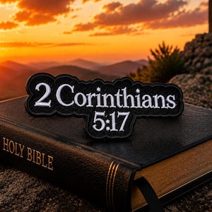 May include: A black leather Holy Bible with gold lettering and a black patch that reads "2 Corinthians 5:17" in white lettering. The Bible is open with a black ribbon bookmark. The background features a sunset over a mountain range.