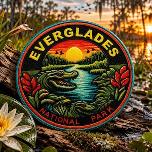May include: Embroidered Everglades National Park patch. The circular patch features a sunset over a river with alligators and the words "EVERGLADES NATIONAL PARK" in yellow lettering. The patch is on a piece of wood.
